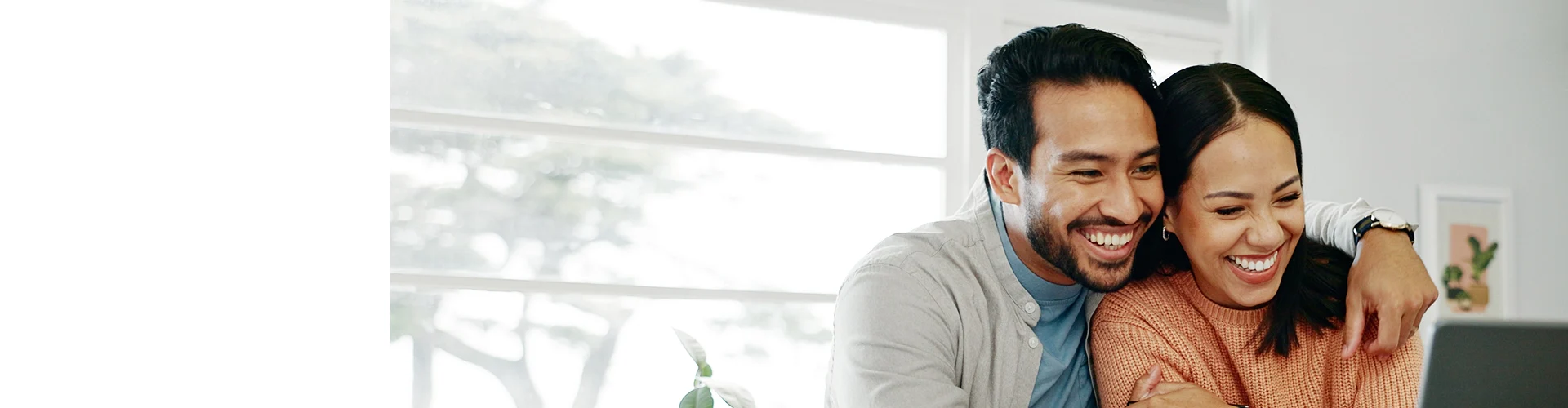 A couple sits close together at a desk, focused on their laptops, with large windows in the background and plants nearby.